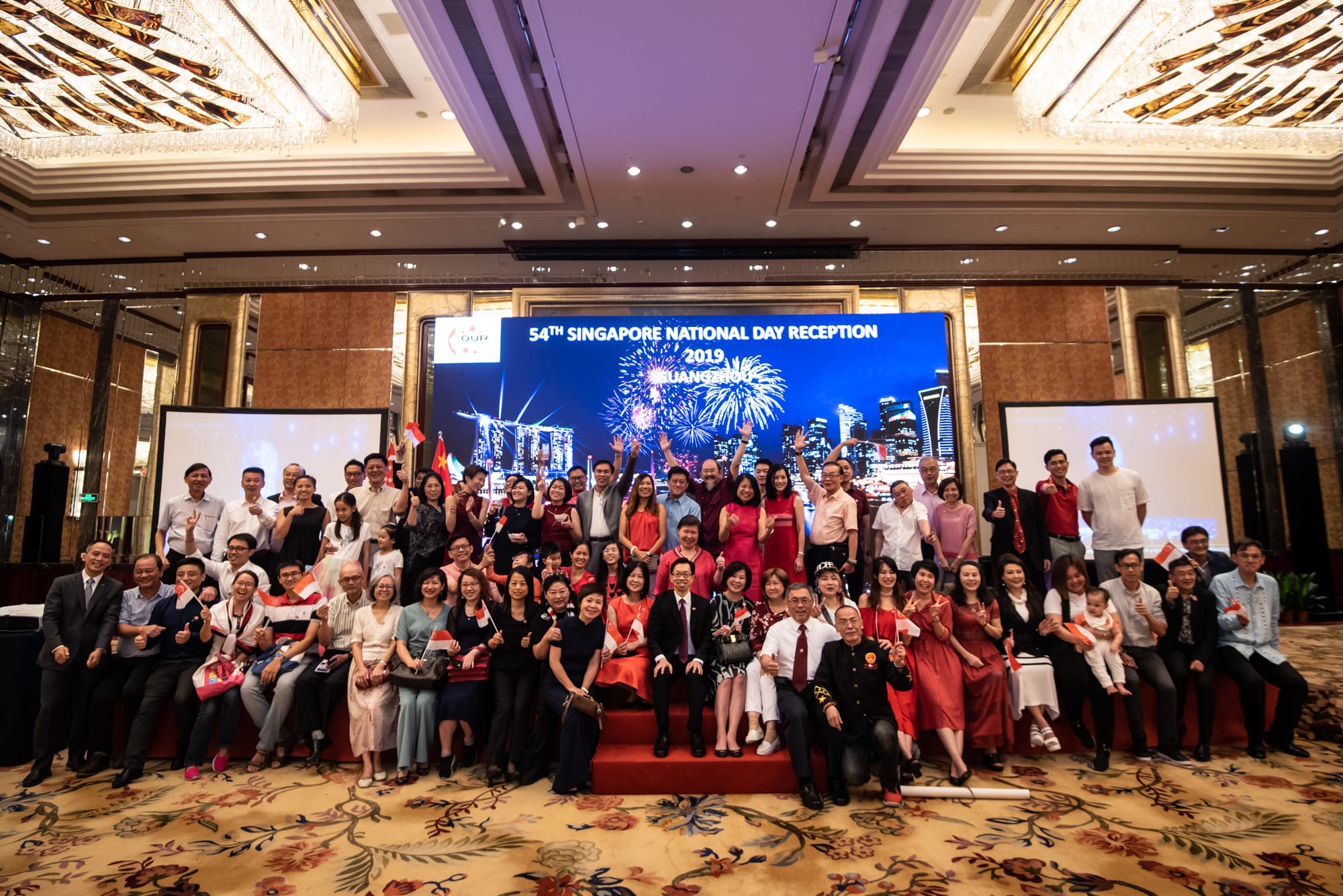 Group poses at 54th Singapore National Day reception with city skyline and fireworks on a screen.